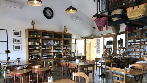 Ein gemütliches Café mit Holztischen und -stühlen, einem großen Bücherregal voller Bücher und Flaschen sowie einer Uhr an der Wand.