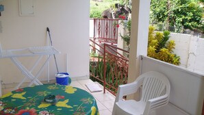 Un petit balcon avec une table recouverte d’une nappe à motifs floraux, une chaise en plastique blanche et un seau bleu.
