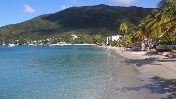 Une plage avec une eau cristalline, des bateaux et une montagne en arrière-plan.