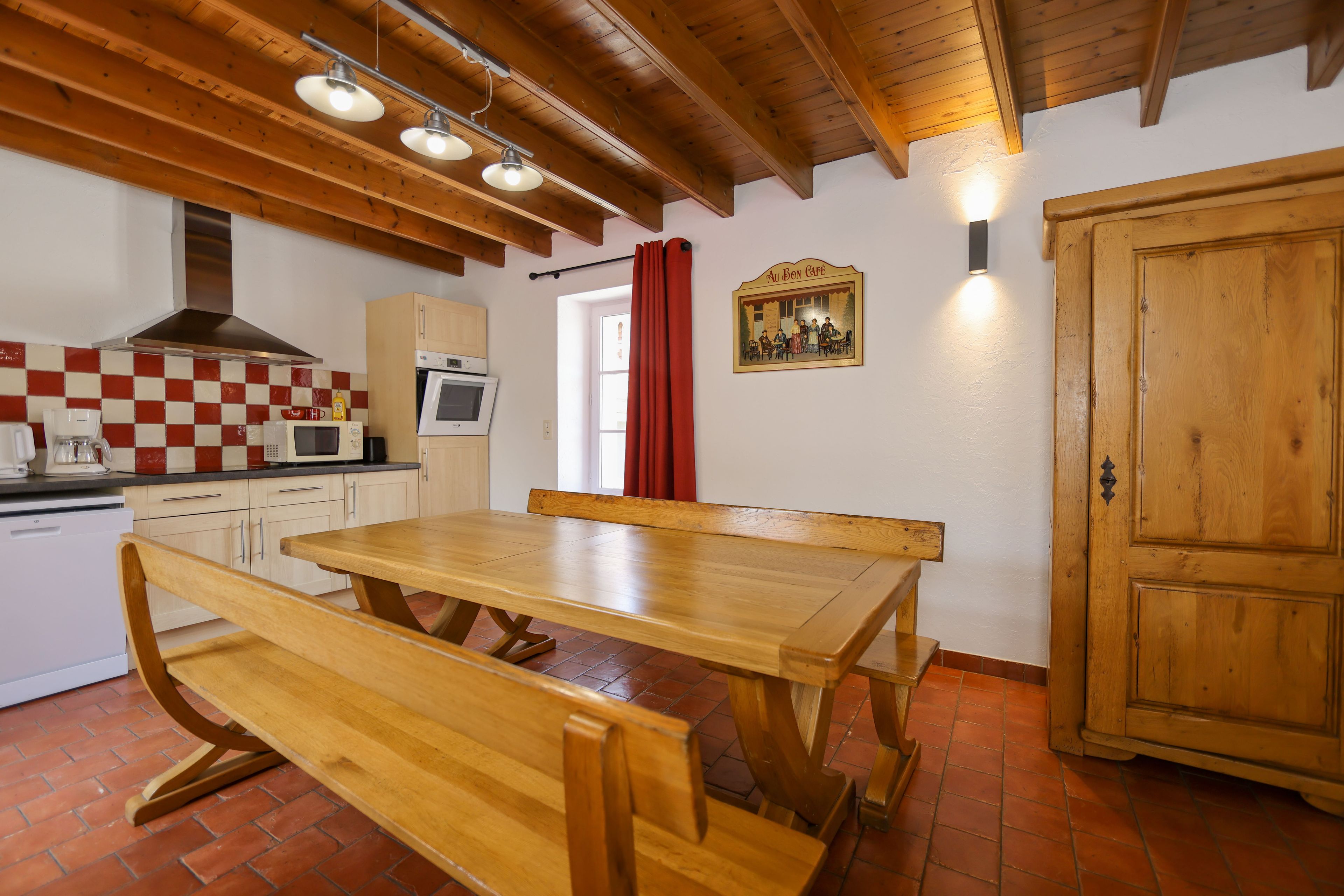 Une cuisine avec des poutres apparentes en bois, un crédence à carreaux, une table en bois avec des bancs, et une porte dotée d’un panneau en bois.