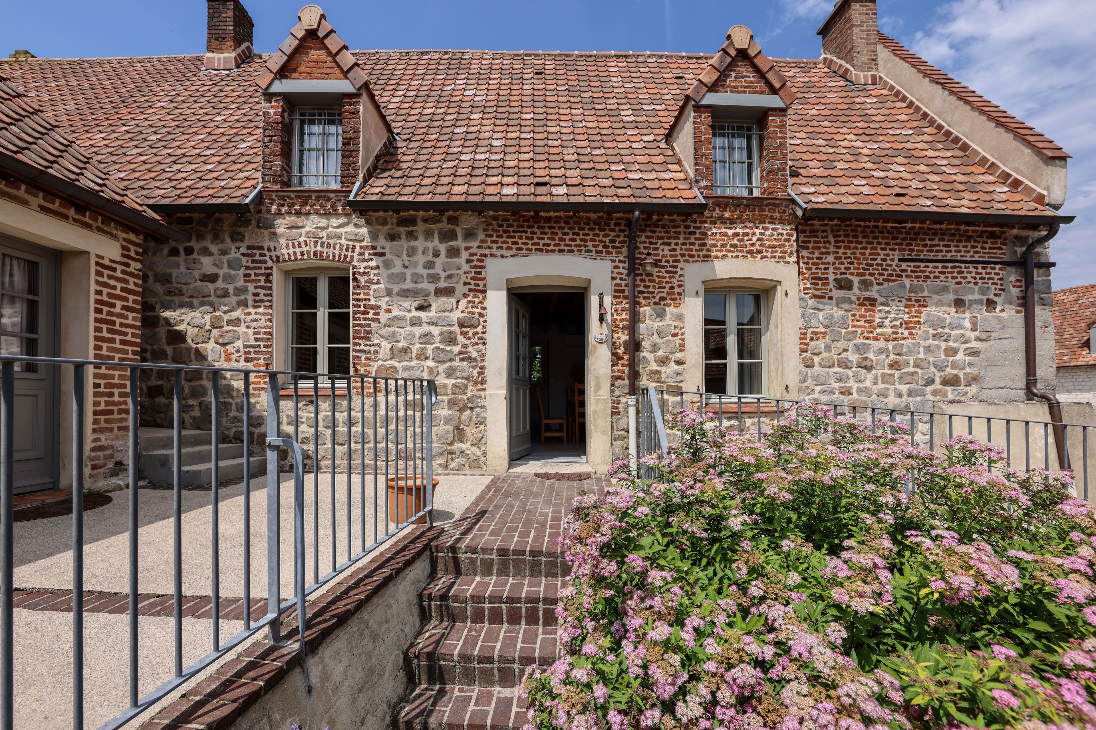 A stone building with a tiled roof, a front entrance, and a small garden with pink flowers.