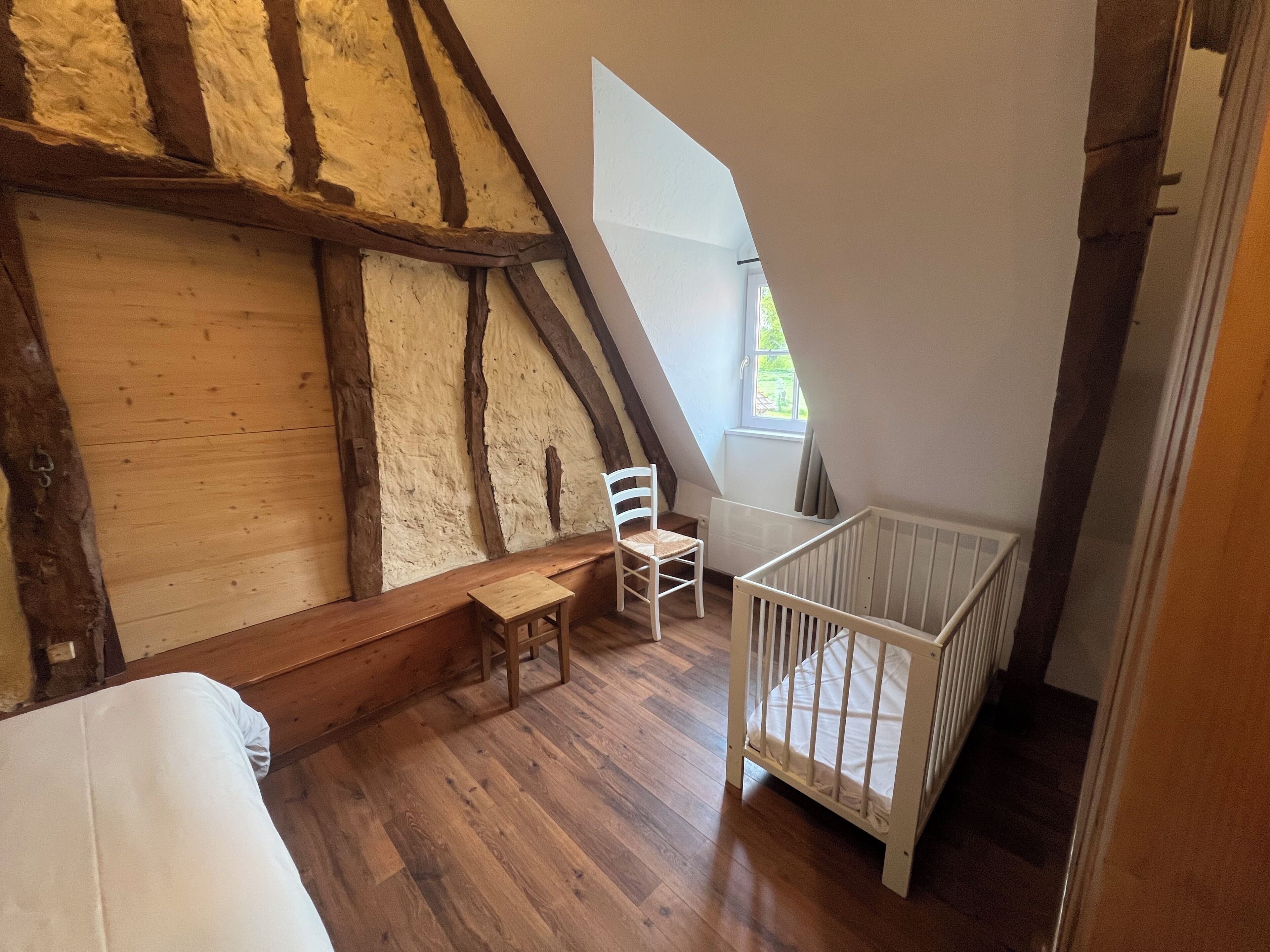 A room with a sloped ceiling, wooden beams, a crib, a chair, and a small table.