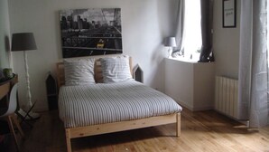 A bedroom with a bed, a cityscape painting, a lamp, and a window with curtains. - Grand T1 in hyper-center in a classified building (Dijon)