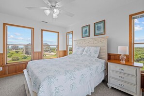 A bedroom with a bed, a nightstand, a lamp, a ceiling fan, and large windows offering a view of houses and greenery. - Soundfront Home w/ Hot Tub, Easy Windsports Access, Sound Boardwalk, and More! (Frisco)