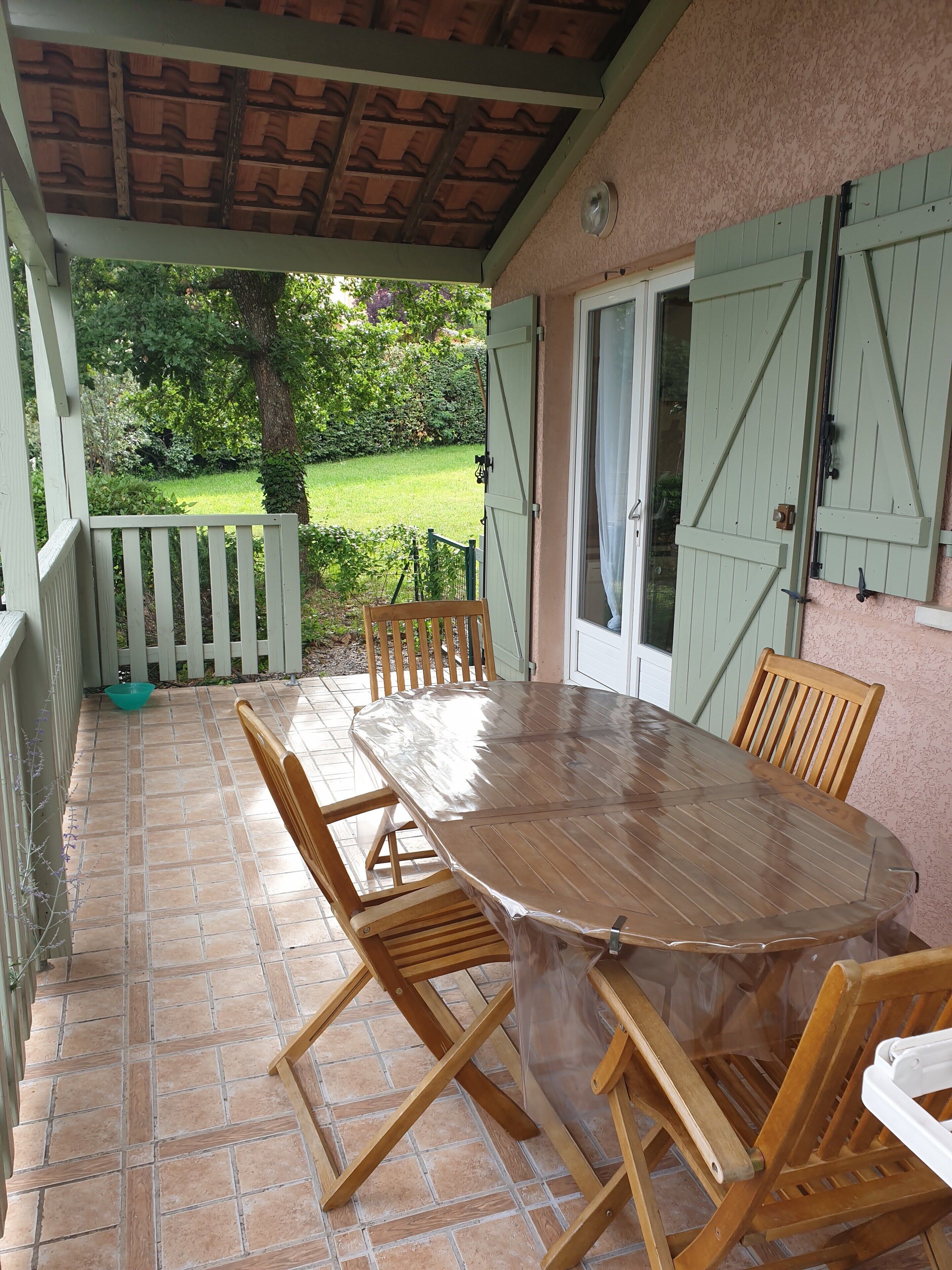 Une terrasse couverte avec une table et des chaises en bois, un bol blanc et une clôture verte en arrière-plan.