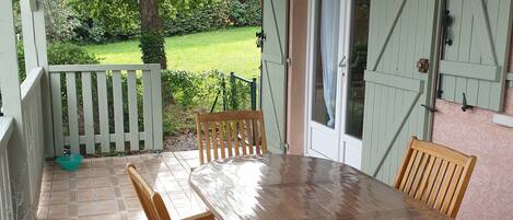 Une terrasse couverte avec une table et des chaises en bois, un bol blanc et une clôture verte en arrière-plan.