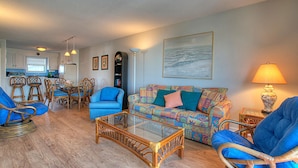 A living room with wicker furniture, a colorful sofa, and a glass coffee table. - ☀️Sands V A3: Oceanfront, Panoramic Views, Shared Pool☀️ (Carolina Beach)