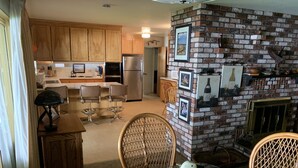 A spacious kitchen with wooden cabinets, a stainless steel refrigerator, and a brick fireplace. There are two wicker chairs and a glass table in the foreground.
