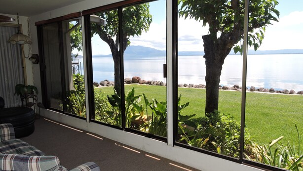 A room with a large sliding glass door leading to a view of a lake and mountains. Inside, there is a plaid sofa and a small table.