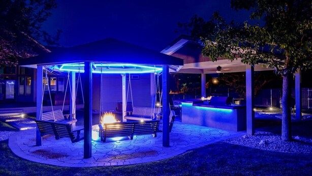 A gazebo with a fire feature and blue ambient lighting, surrounded by a stone patio and a pool area. - Claim Jumpers Cottage (Baker City)