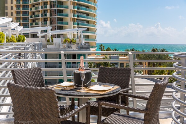 A balcony with wicker chairs and a table, overlooking a pool and a building with a view of the ocean.