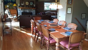 Un coin repas avec une table en bois dressée pour six personnes, entourée de chaises en osier ornées de coussins roses. Une cheminée en pierre avec une cheminée décorative se trouve au fond, et un canapé est visible dans le salon attenant.