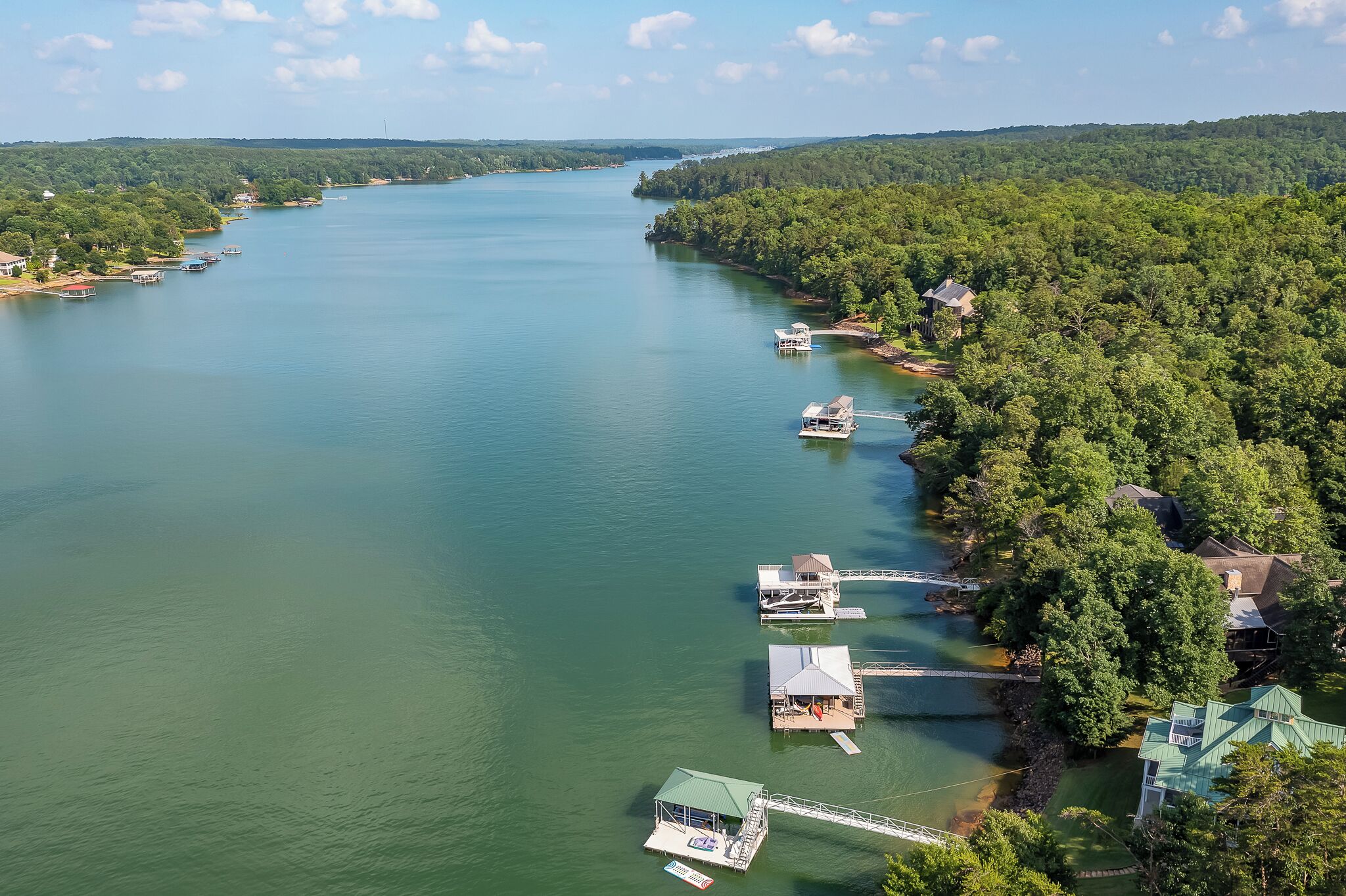 Vista aérea de uma propriedade à beira do lago, com várias docas e barcos, rodeada por uma floresta densa.