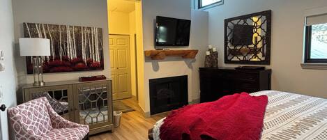 Une chambre à coucher comprenant un lit, une télévision, une cheminée, un fauteuil et une porte donnant sur une autre pièce.