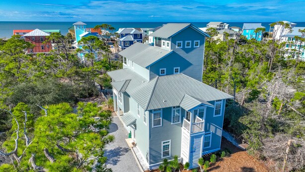 Un'ampia casa a due piani con tetto grigio e rivestimento blu, circondata da pini e vista libera sull'oceano.
