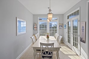 A dining area with a white table and chairs, a chandelier, and large windows with a view.