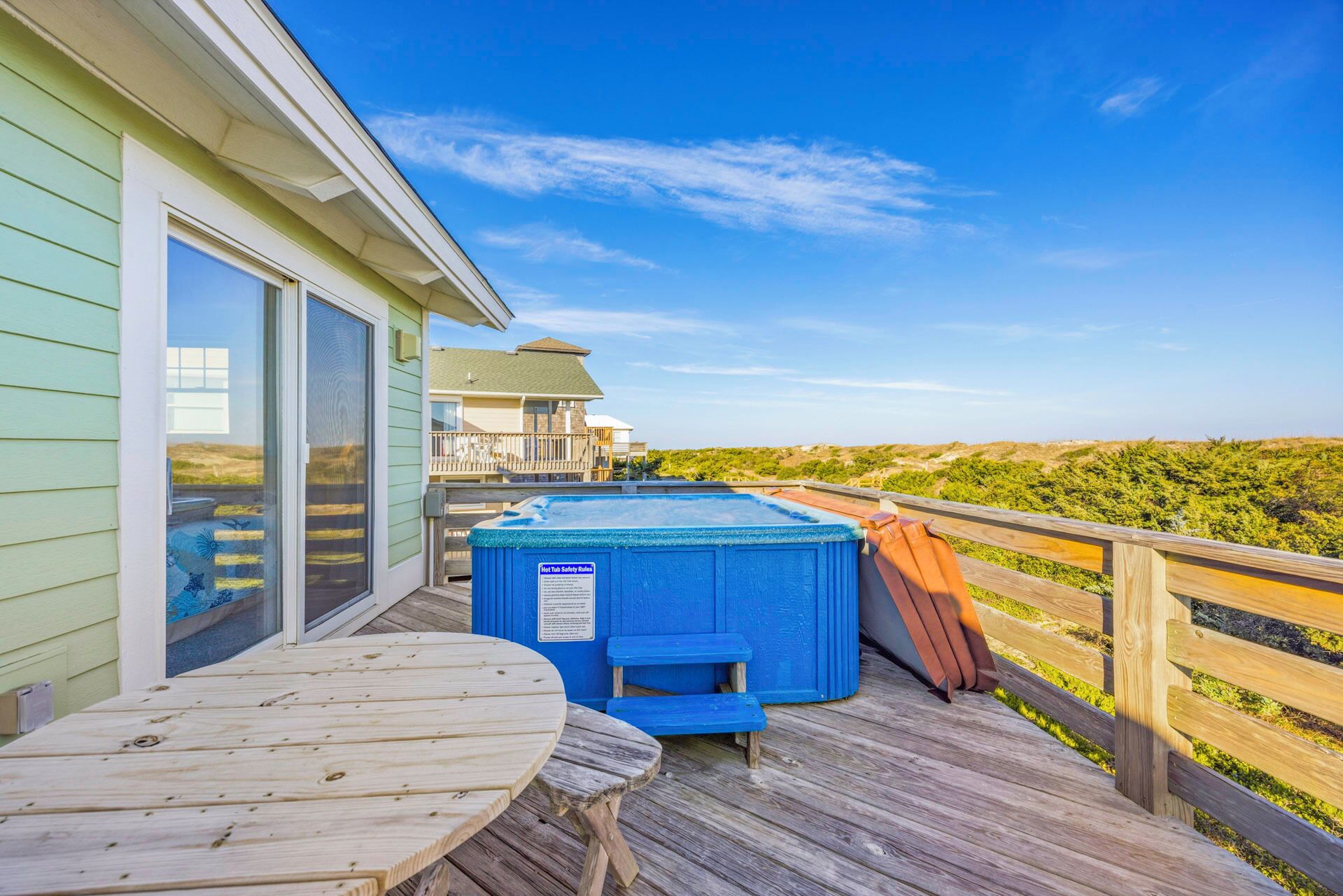A wooden deck with a hot tub, a table, and a view of a beach and distant buildings.