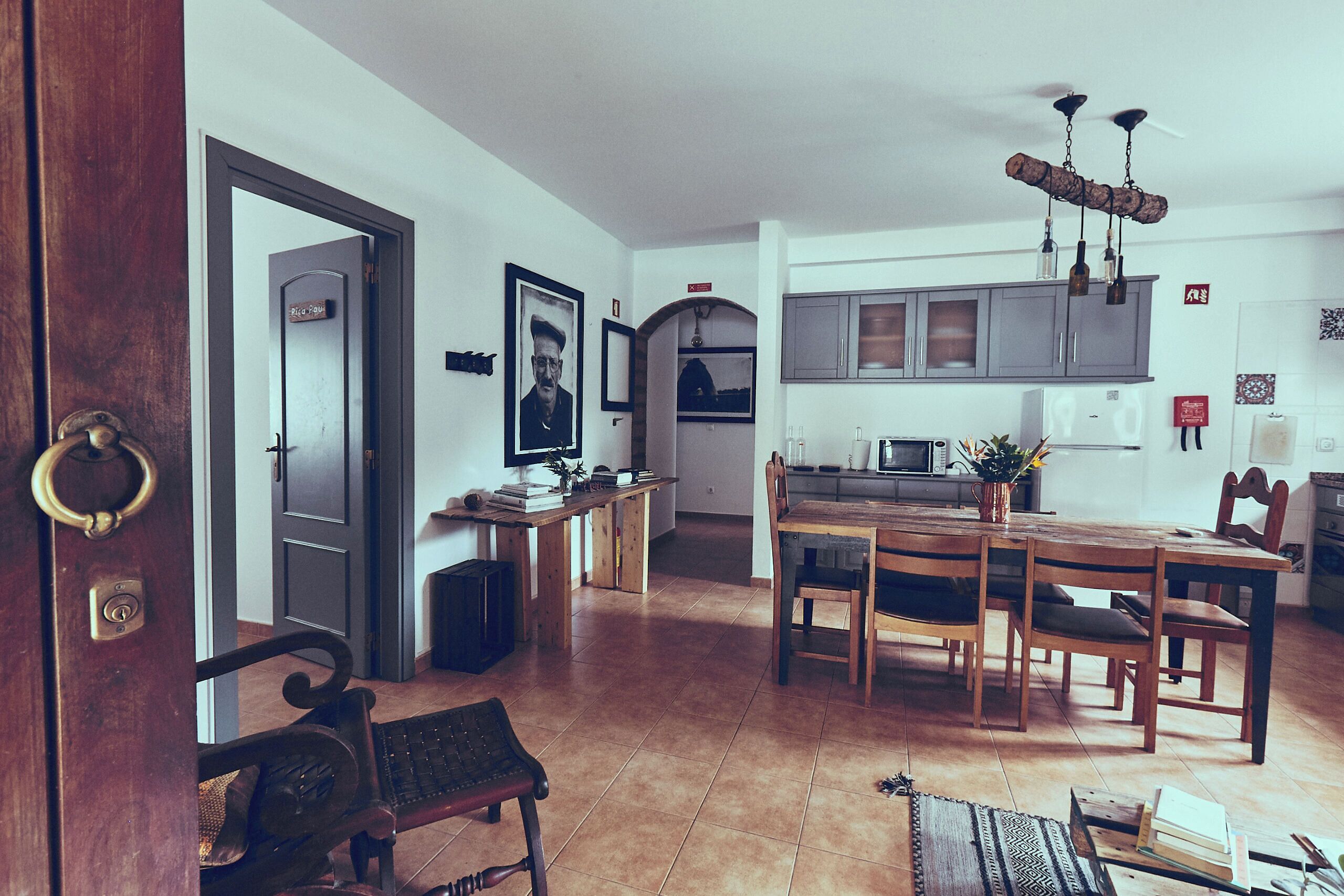 Un amplio salón con mesa de madera, sillas y un banco. Se ve una cocina con armarios grises y una lámpara colgante al fondo.