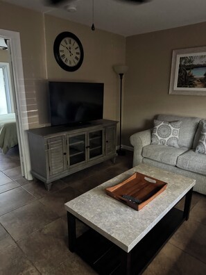 A living room with a television on a cabinet, a coffee table with a tray, a sofa, and a clock on the wall.