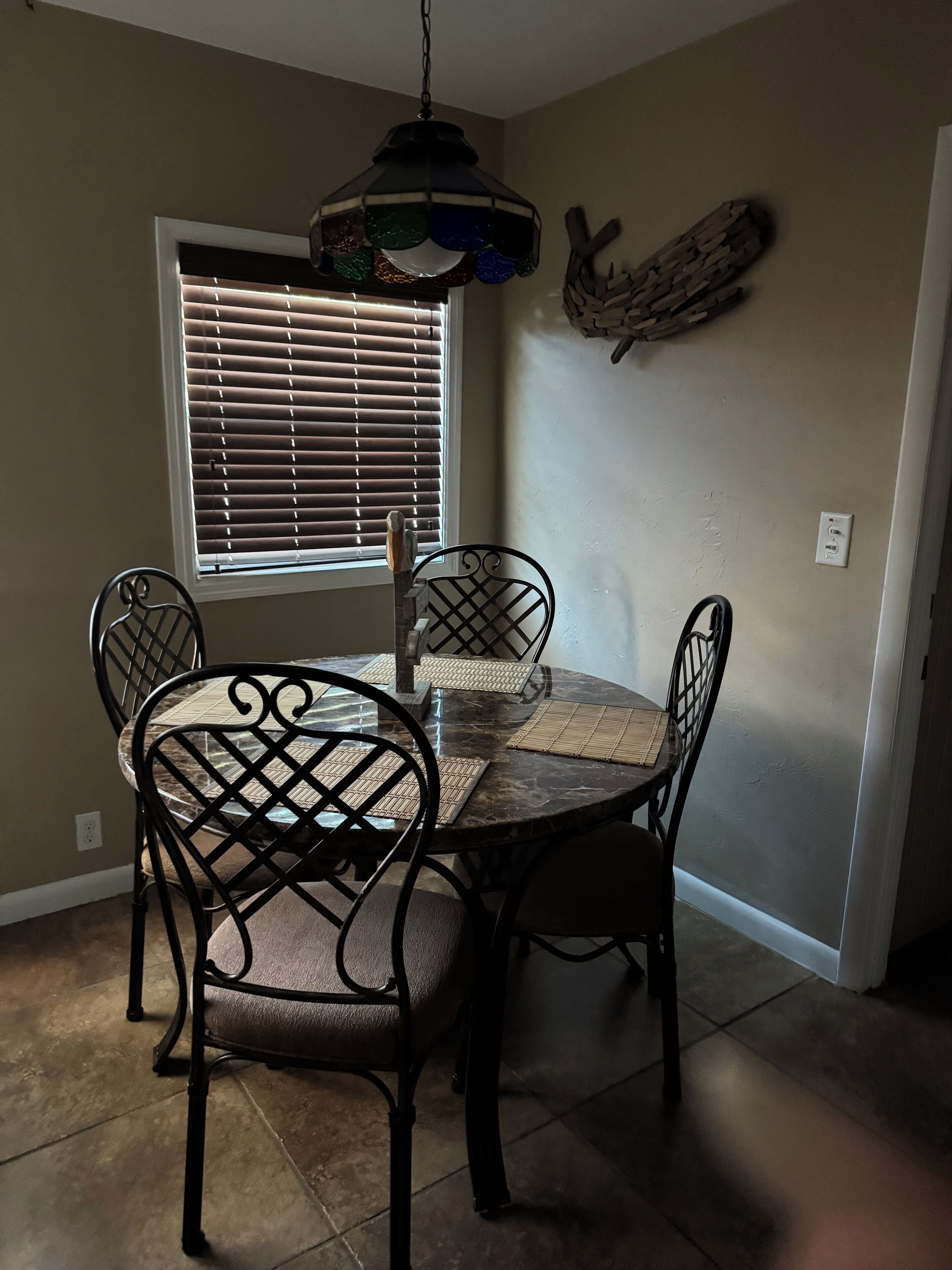 Un coin repas comprenant une table ronde, quatre chaises, un luminaire suspendu, une fenêtre avec des stores et un élément décoratif fixé au mur.