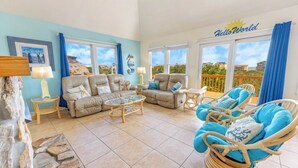 A spacious living room with a beige sofa, a glass coffee table, and wicker chairs. There are two windows with blue curtains, a stone wall, and a 'Hello World' sign above the windows.