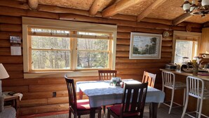Interior acogedor de una cabaña con detalles de madera, una mesa de comedor con sillas, una zona de cocina y ventanas amplias que dan al bosque.