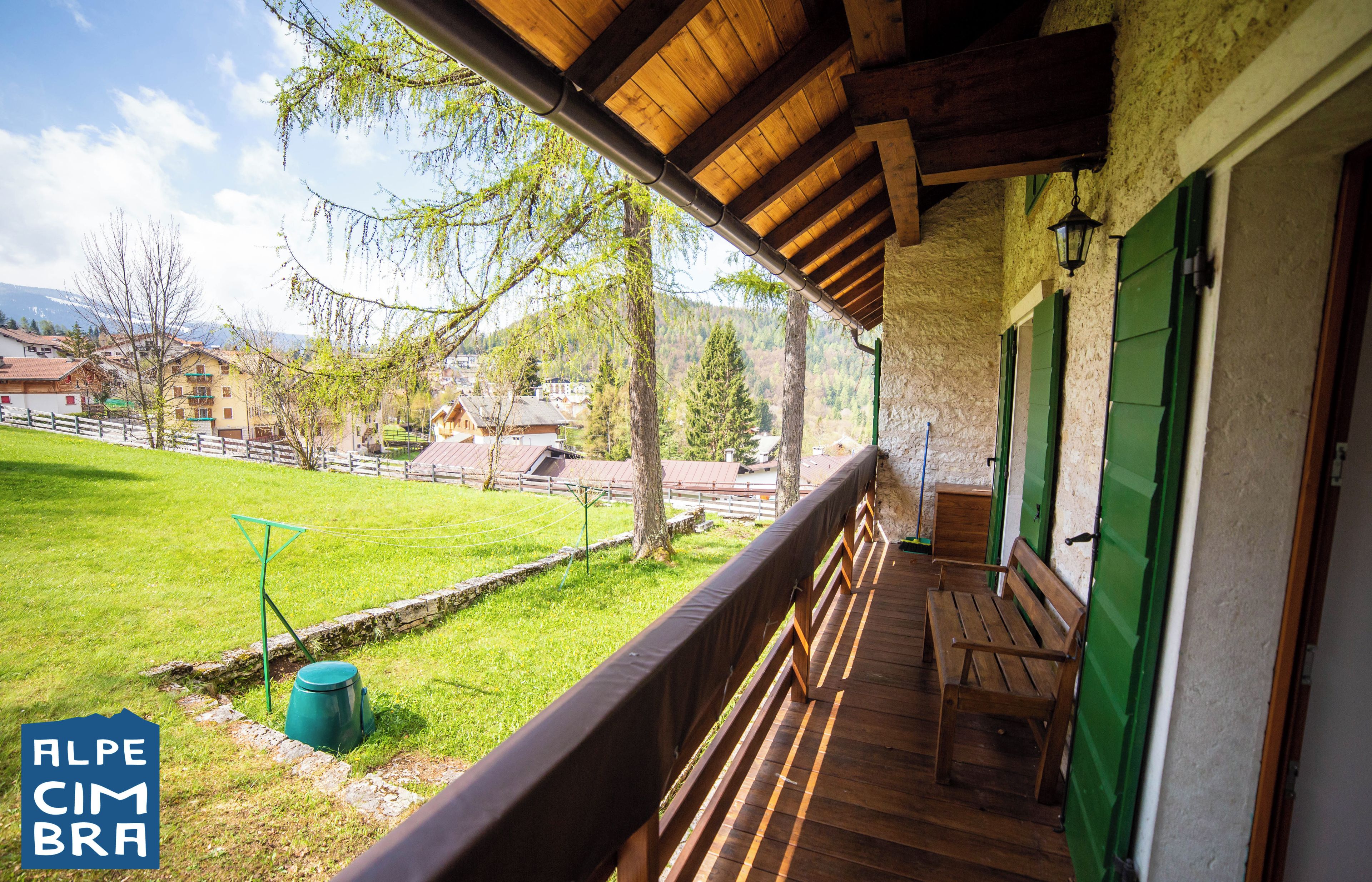 A wooden balcony with a bench, a green bucket, and a view of a green field and buildings in the background.