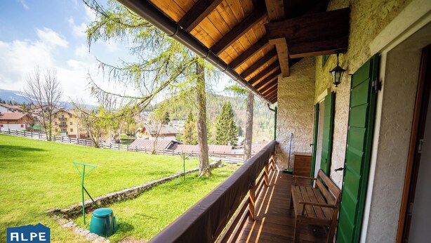 Un balcón de madera con un banco, un cubo verde y vistas a un campo verde y edificios al fondo.