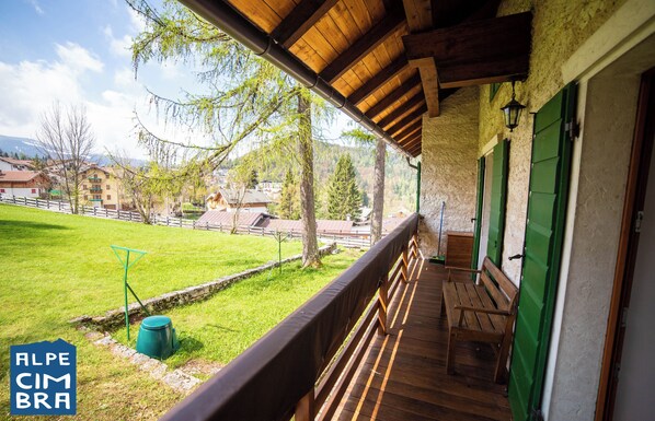Un balcon en bois avec un banc, un seau vert et une vue sur un champ verdoyant et des bâtiments en arrière-plan.
