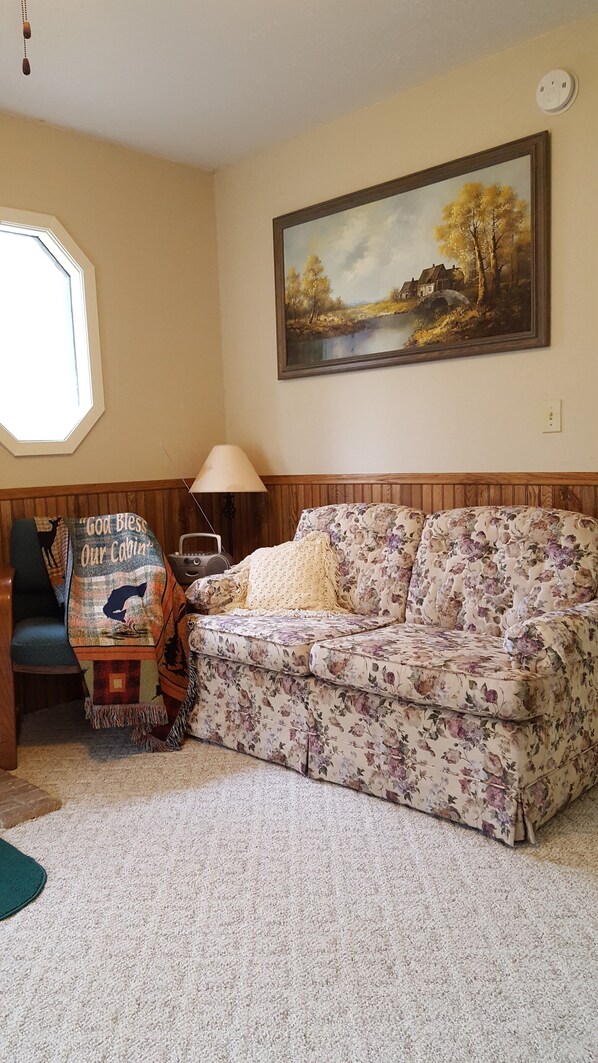 Una sala de estar acogedora con un sofá floral, un cuadro de un paisaje y una ventana con persianas.