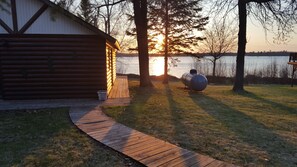 Una cabaña de troncos con una terraza de madera que da a un lago, rodeada de árboles y un cielo despejado.