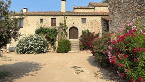 Un edificio rústico de piedra con un arco central, una chimenea y un pequeño jardín con plantas en flor.