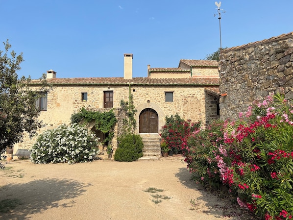 A rustic stone building with a central arched doorway, a chimney, and a small garden with flowering plants.