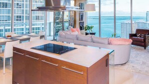 A modern kitchen with a large island, dining area with white chairs, and a sofa with a view of the ocean and buildings. - Prime Brickell Suite | Walk to City Center (Miami)