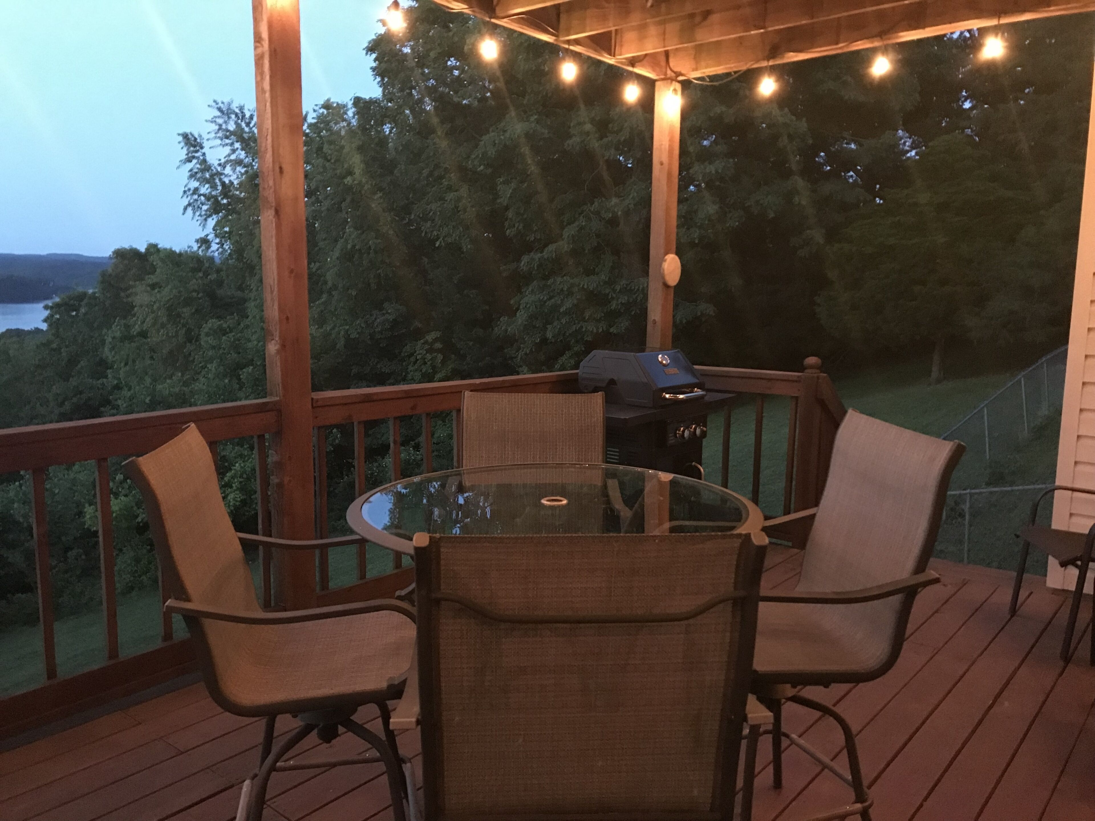 A covered outdoor deck with a glass table surrounded by chairs, a barbecue grill, and string lights.