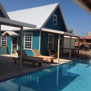 A poolside area with lounge chairs and a house with a blue exterior and white trim.