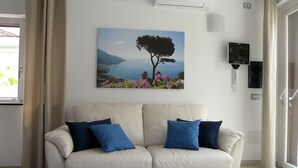 Una sala de estar con un sofá color beige, cojines azules, un cuadro de un árbol junto al mar y un aire acondicionado instalado en la pared.