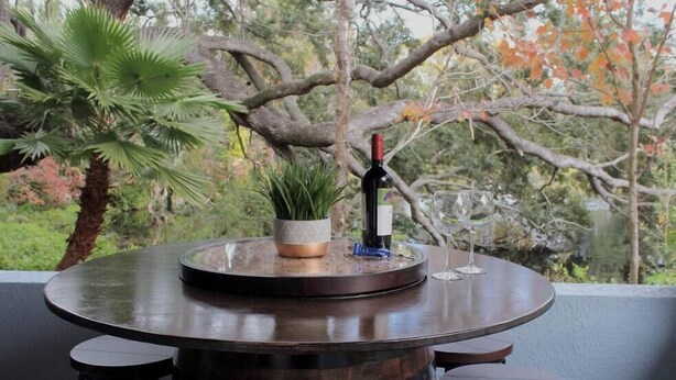 A round wooden table with a wine bottle and glass, surrounded by stools, set against a backdrop of trees and foliage.