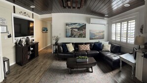 A cozy living room with a dark leather sofa, a coffee table, and a TV. There are decorative pillows, a vase with flowers, and artwork on the walls.