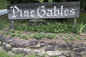 Uma placa de madeira com o texto 'Pine Cables' emoldurada por um muro de pedra e vegetação.