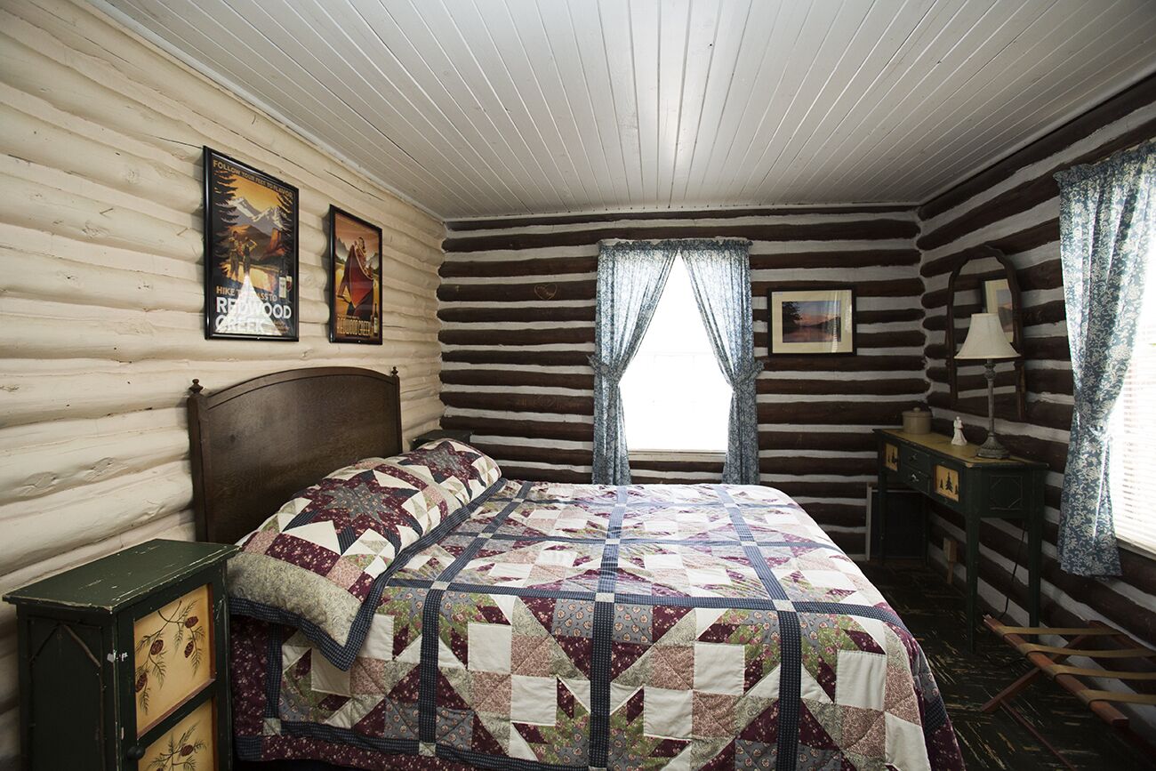 A log cabin room with a wooden bed, a nightstand, and a window with curtains.