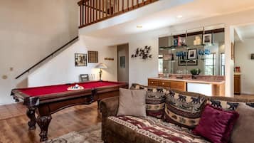 A living room with a pool table, a sofa with patterned cushions, and a kitchen area visible through an open door.