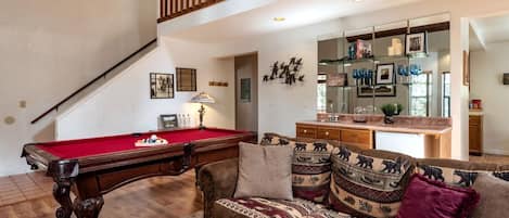 Una sala de estar con mesa de billar, sofá con cojines estampados, y un área de cocina visible a través de una puerta abierta.