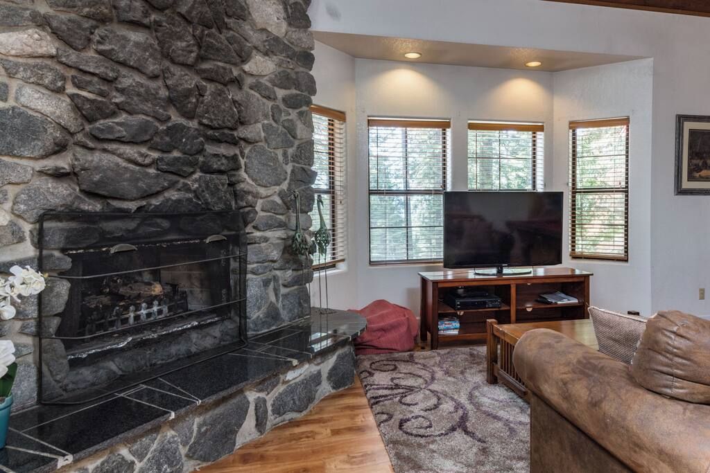 A living room with a stone fireplace, a flat-screen TV, and a large window with blinds.