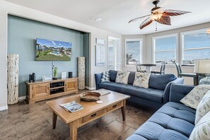 A living room with a blue sofa, wooden coffee table, and a television mounted on the wall. - A Shore Thing - Overlooking Pool/ Ocean Views to South/ Bay View to North (Port Aransas)