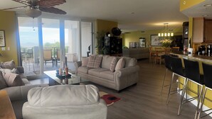 A spacious living area with a sofa, a coffee table, and a dining area with chairs and a bar. - Luxurious, Newer Oceanfront, Professionally Decorated, Beautiful Condo (Cocoa Beach)