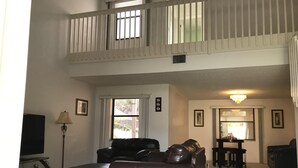 Un salón espacioso con techo alto, un balcón con barandilla y una zona de comedor con mesa y sillas.