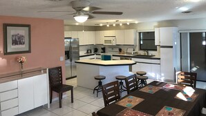 Una cocina moderna con armarios blancos, una isla central con taburetes, un ventilador de techo y un comedor con mesa y sillas.