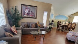 Ein geräumiges Wohnzimmer mit Holzboden, einem grauen Sofa, einem Glastisch und einem Essbereich mit Holzsesseln und einem Tisch.
