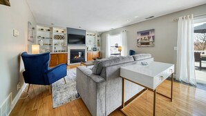 Un salon moderne avec un canapé d’angle gris, une table blanche, un fauteuil bleu, une cheminée et un téléviseur.